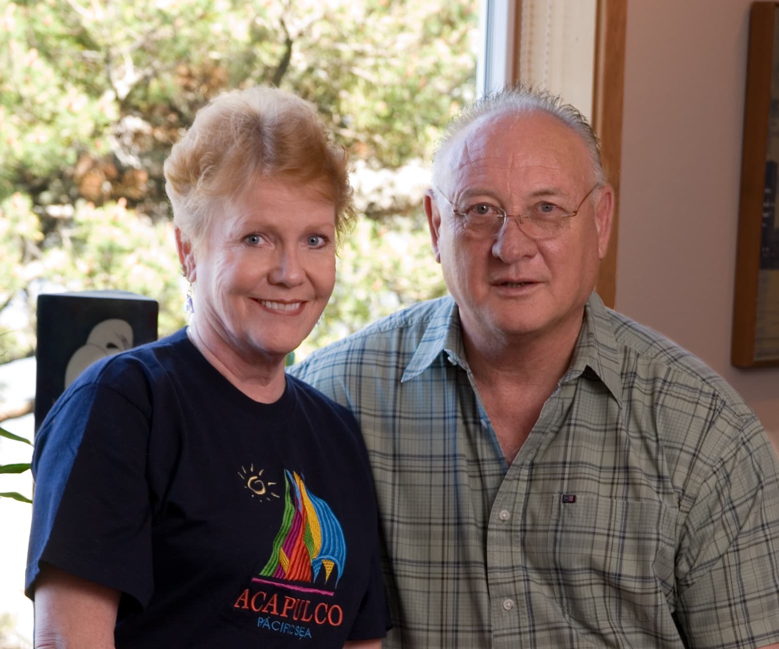 A woman with short blonde hair and a man with glasses pose together in front of a window.