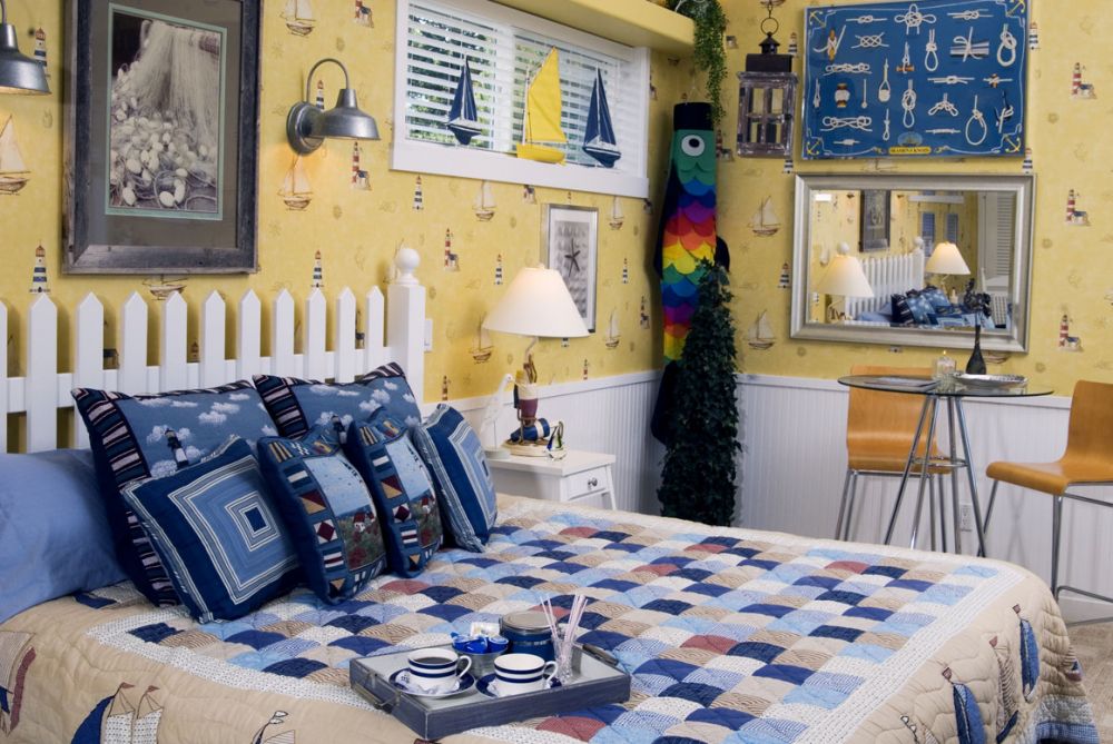 Bedroom with a white picket fence headboard, blue and white patchwork quilt, nautical themed artwork, yellow wallpaper, and a small table with chairs.