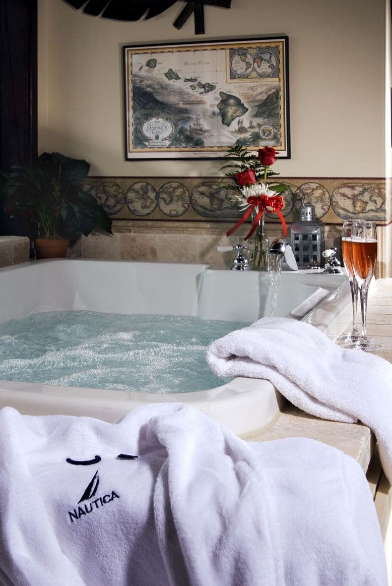 Close-up of a bubbling jacuzzi tub with white plush robes, two wine glasses, and a floral arrangement.