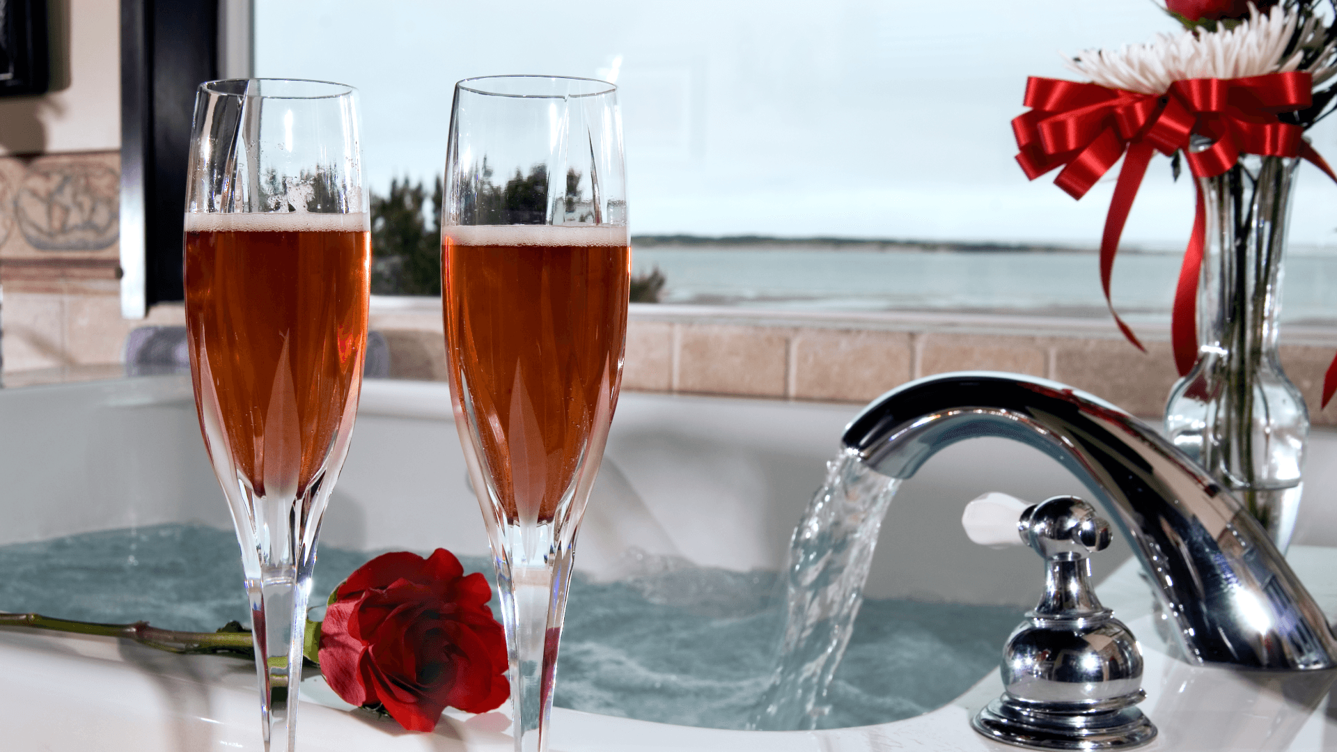 Two champagne flutes filled with rose-colored bubbly sit by a bubbling hot tub, alongside a single red rose and a decorative ribbon in the background.