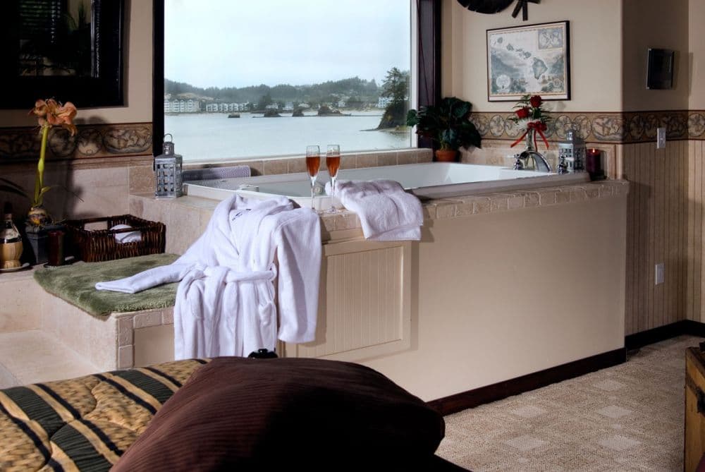 Bathroom with a built-in Jacuzzi tub, white robes, two wine glasses, and a large window overlooking the waterfront.