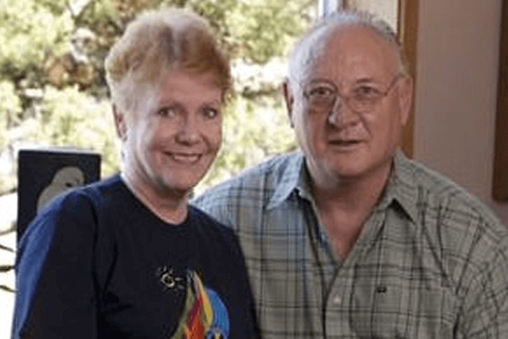 A woman with short blonde hair and a man in a plaid shirt smile for the camera indoors.