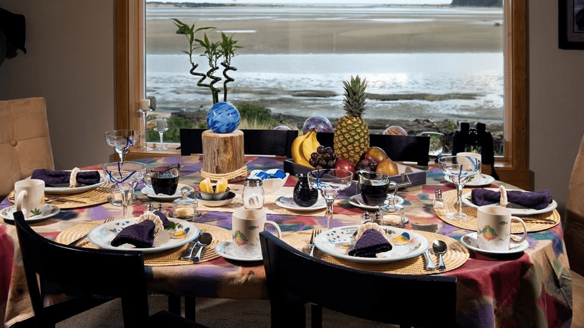 A beautifully set dining table with colorful dishes and fruits, overlooking a serene landscape.