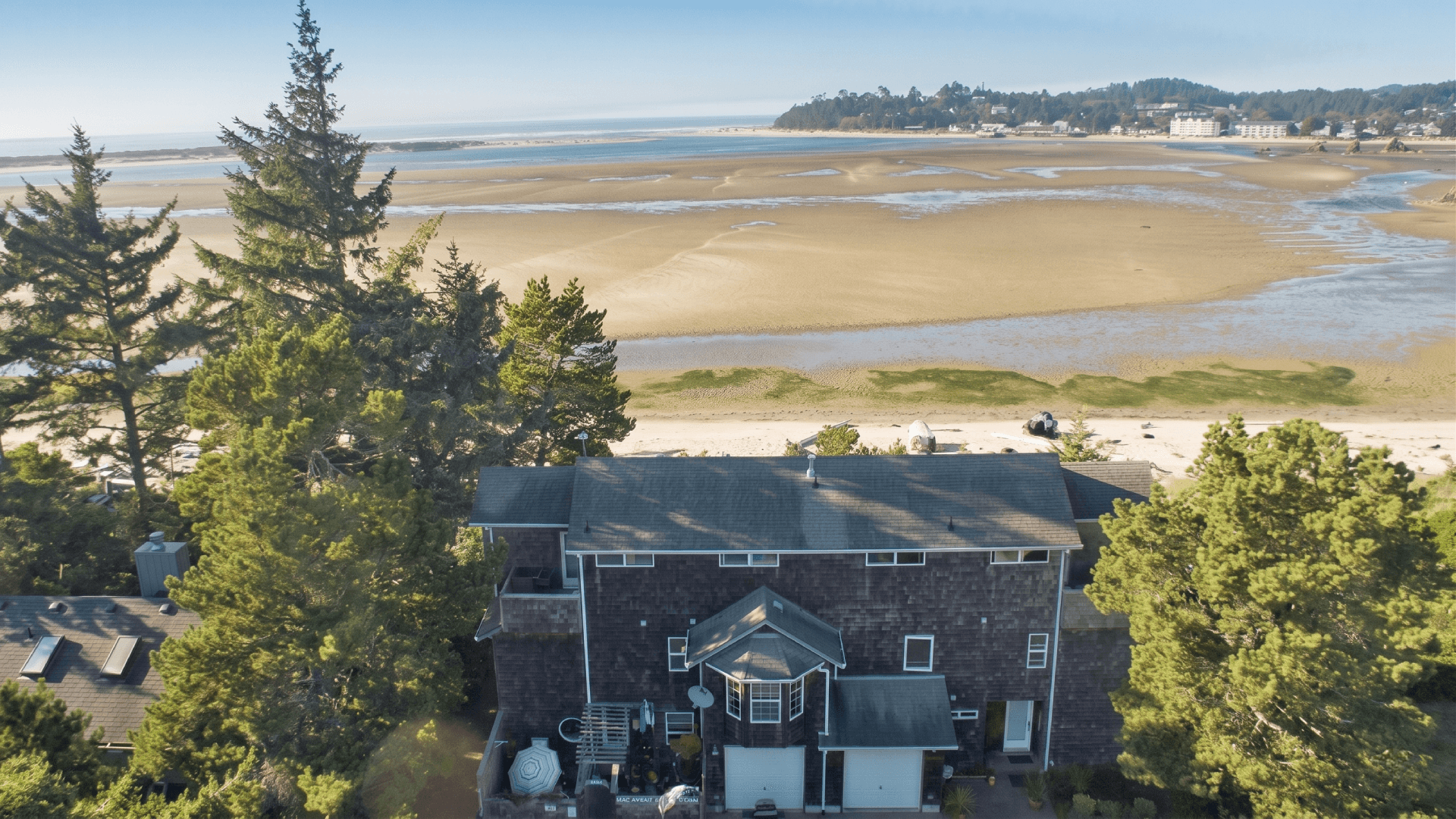 Aerial view of a coastal house surrounded by trees, with a sandy beach and tidal flats in the background.
