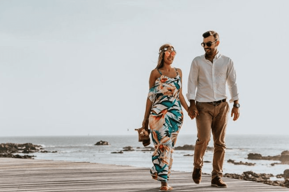 A couple walks hand in hand along a wooden boardwalk by the ocean.