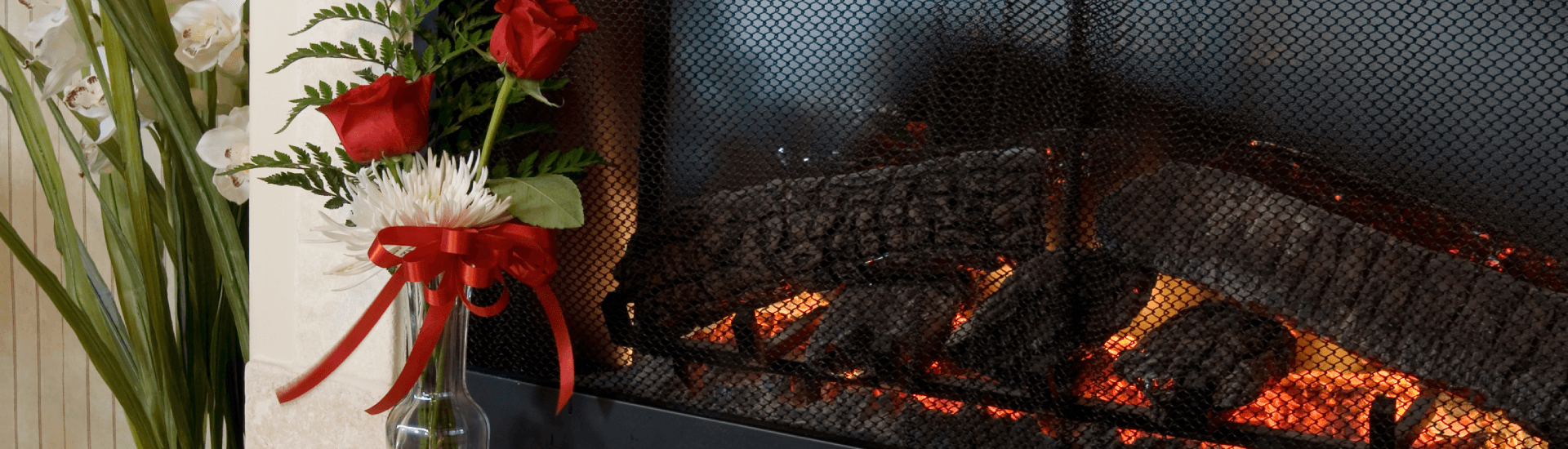 A cozy fireplace adorned with logs and flames, next to a vase of red roses and greenery.